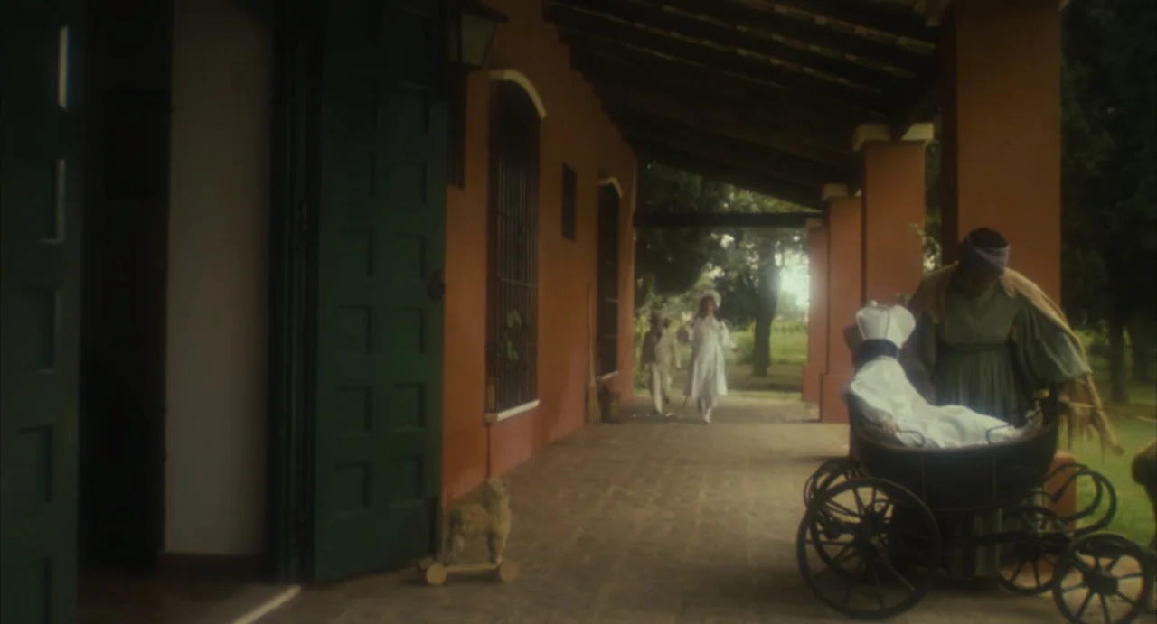 Camila (1984) movie still: wide — A long terracotta-colored porch with dark green doors and stone flooring overlooks a yard. A woman pushes a…