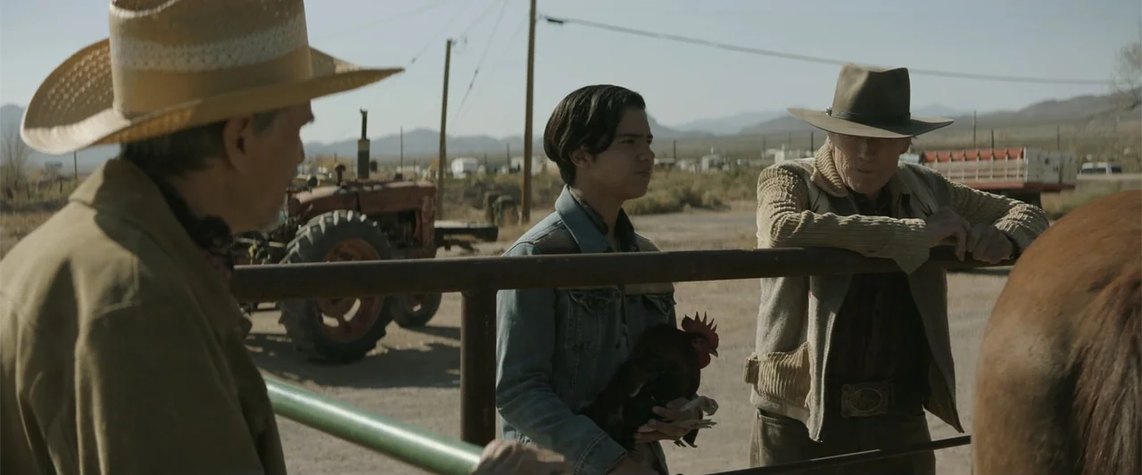 Cry Macho (2021) movie still: medium — An arid, open desert environment features a metal pipe gate and a rusty tractor. Three males converse…