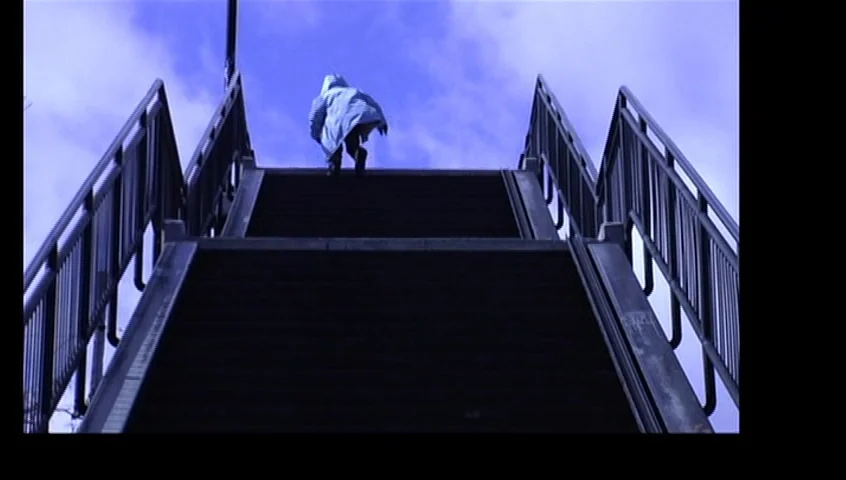 The Tracey Fragments (2007) movie still: wide — A metal pedestrian overpass rises towards a blue cloudy sky. A single hooded figure walks up the…