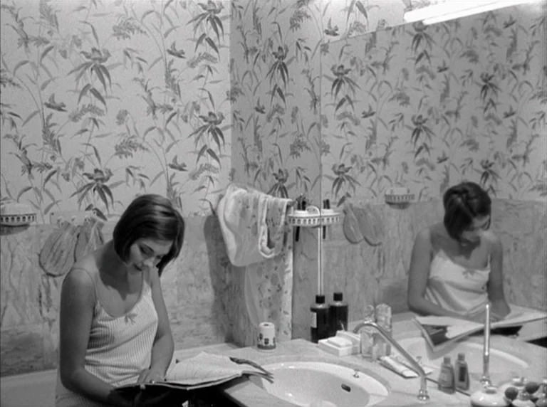 Une Femme Mariée (1964) movie still: medium — A bathroom features patterned floral wallpaper and a sink with a wall-mounted mirror. A woman sits on…
