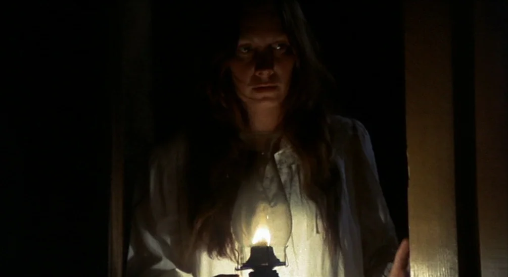 The Hired Hand (1971) movie still: medium — A woman stands in a pitch-black corridor, illuminated only by a small handheld lantern. She holds the…