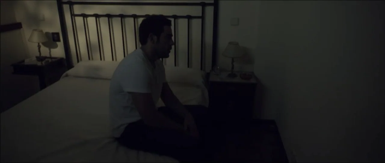 Otro verano (2013) movie still: medium — A dimly lit bedroom features a metal bed frame and two small bedside lamps. A man sits alone on the edge of…