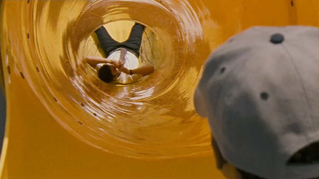 Half Nelson (2006) movie still: medium — A view through a bright yellow plastic tube at a playground. A man lies flat on his back inside the slide…