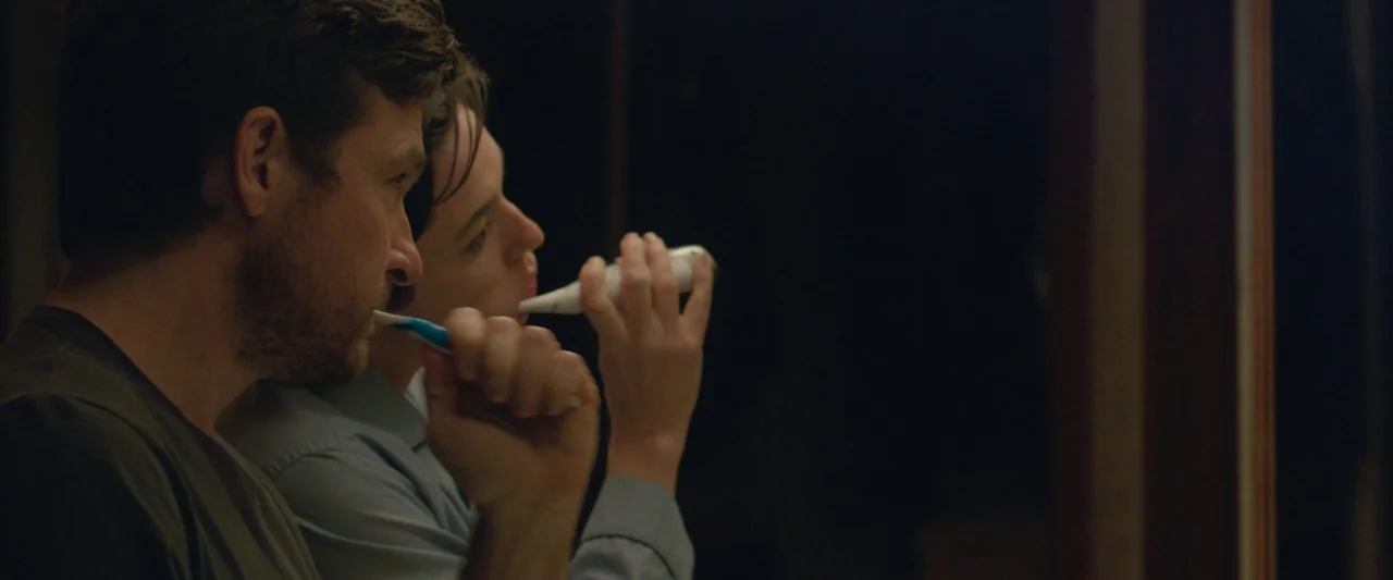 The Gift (2015) movie still: medium — A dark bathroom interior features two people standing side by side in profile. They both hold toothbrushes to…