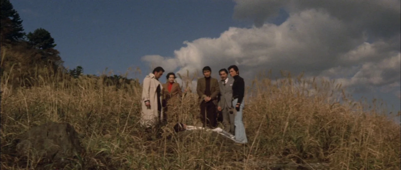 Terror of Mechagodzilla (1975) movie still: wide — A group of five people stand on a grassy hillside covered in dry weeds. They observe a body lying…