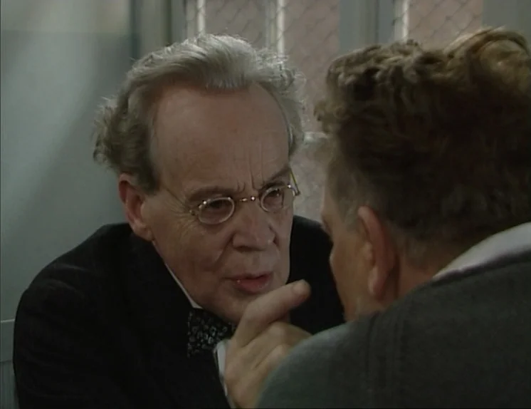 In The Presence of a Clown (1997) movie still: closeup — An interior room features gray walls and a chain-link partition in the background. An older…