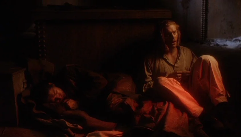 Edward II (1991) movie still: medium — A dark, cramped stone prison cell with a thick, heavy ledge in the background. A man sits awake on the floor…