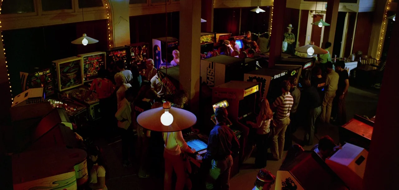 Tron (1982) movie still: wide — A crowded 1980s arcade filled with rows of illuminated upright video games and pinball machines. Multiple people…