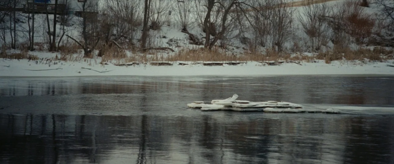 For Ellen (2012) movie still: wide — A dark river flowing past a snow-covered bank with bare, leafless trees. A small cluster of ice floes drifts…