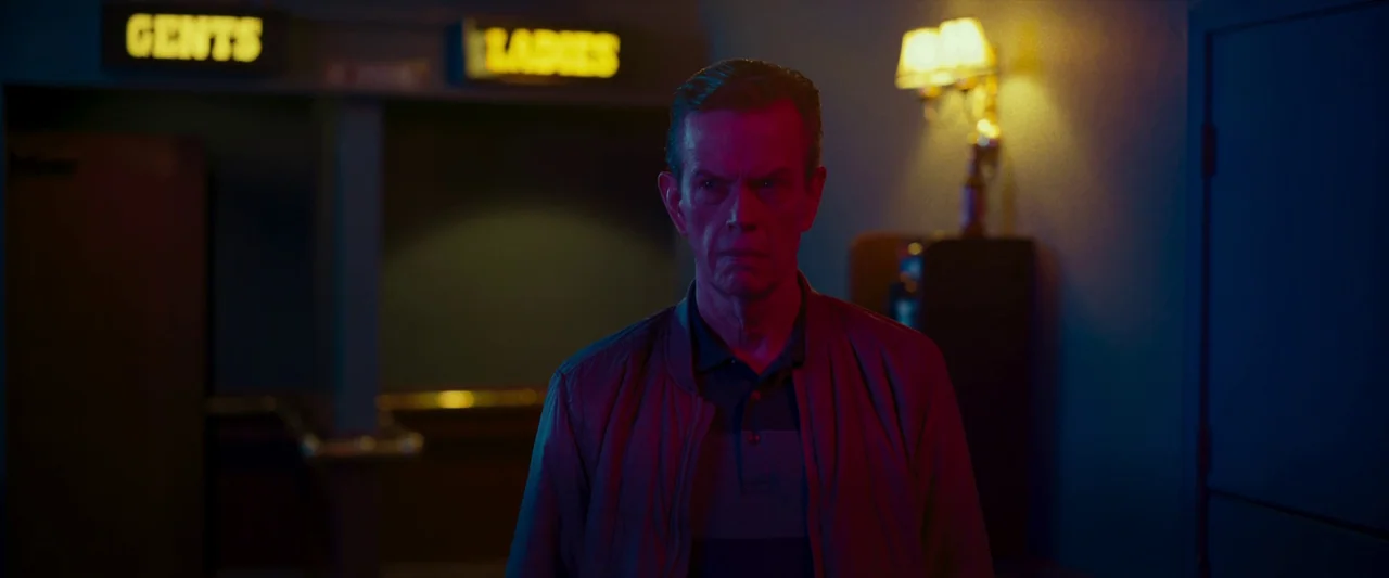 LaRoy, Texas (2023) movie still: medium — A dimly lit indoor hallway features illuminated restroom signs in the background. A middle-aged man stands…