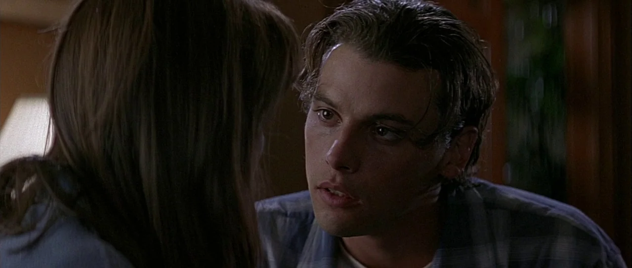 Scream (1996) movie still: closeup — A dark indoor space with wooden furniture visible in the background. A young man stares intently at a woman…