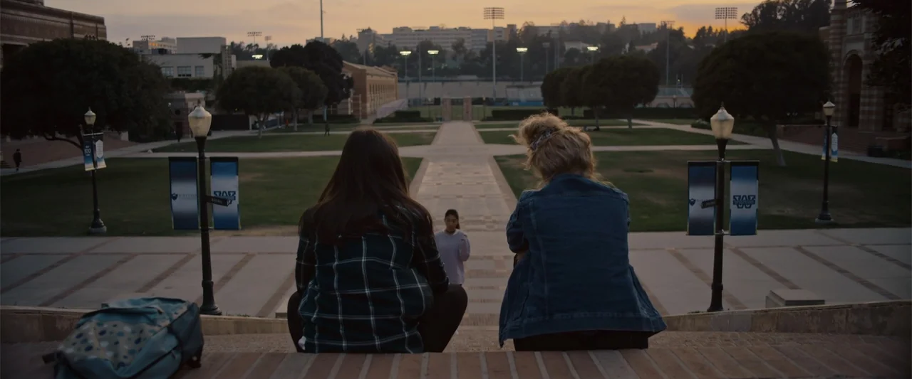 Init!ation (2021) movie still: wide — Two students sit on outdoor stone steps of a university plaza during golden hour. They face a distant third…