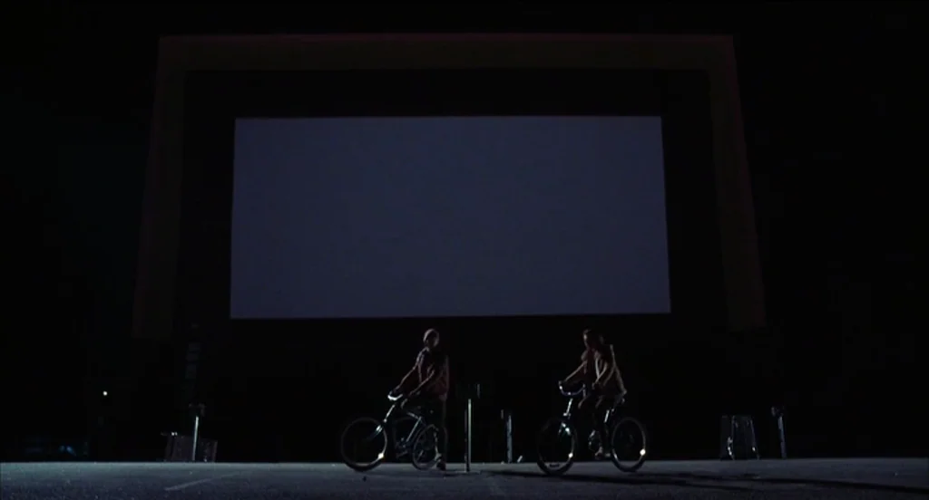 Mysterious Skin (2004) movie still: wide — A vast, dark expanse featuring a large blank projection screen in the background. Two people ride…