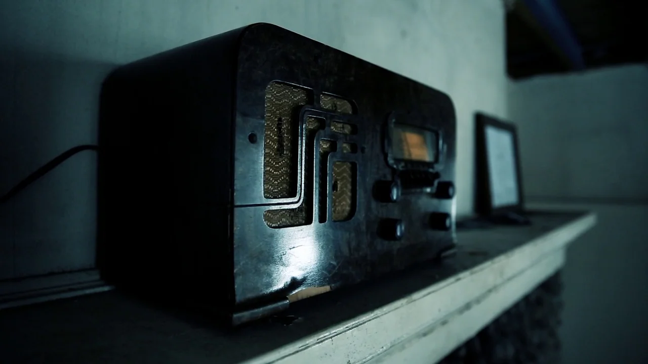 The Deeper You Dig (2019) movie still: closeup — An antique dark wood radio rests on a white mantelpiece against a plain wall. A small framed…