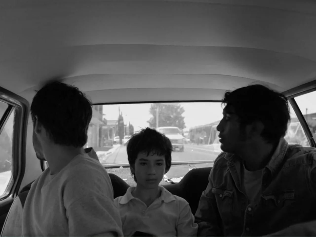 Güeros (2014) movie still: medium — Three individuals sit on the backseat of a car traveling during the day. The camera captures the three subjects…