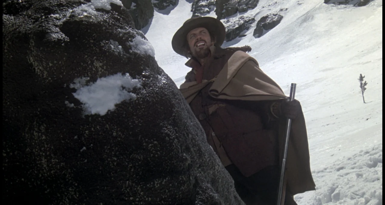 The White Buffalo (1977) movie still: medium — A steep snowy mountain slope with a massive dark boulder in the foreground. A man wearing a felt hat…