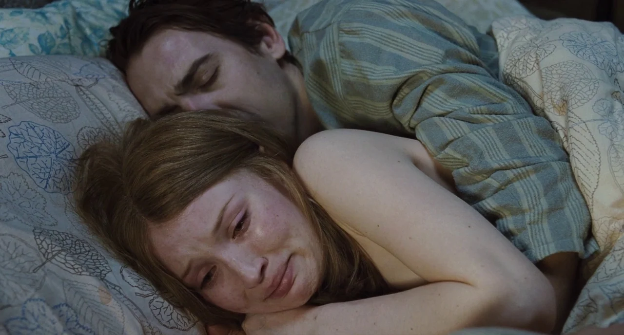 Sleeping Beauty (2011) movie still: medium — A couple lies in bed under patterned linens with leaves printed on the fabric. The man embraces the…