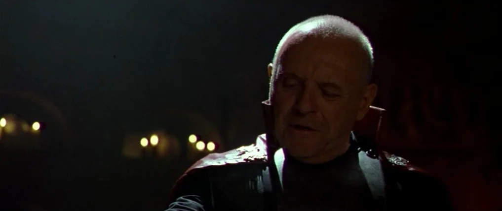Titus (1999) movie still: closeup — A dark, dimly lit stone interior features faint candle glow against distant rounded arches. An elderly man with…