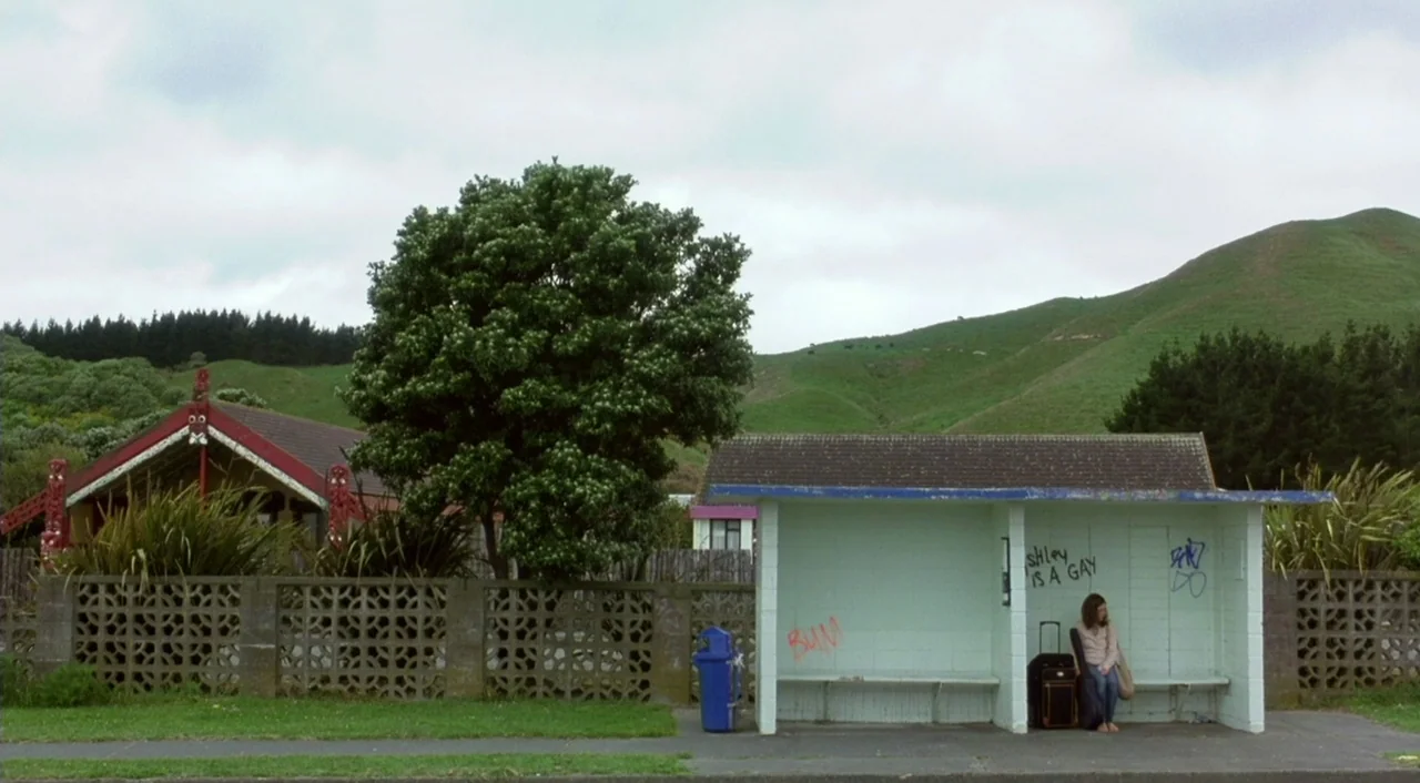 Eagle Vs. Shark movie still: wide — A roadside bus stop sits before a rural landscape with rolling green hills. A single young woman waits alone on…
