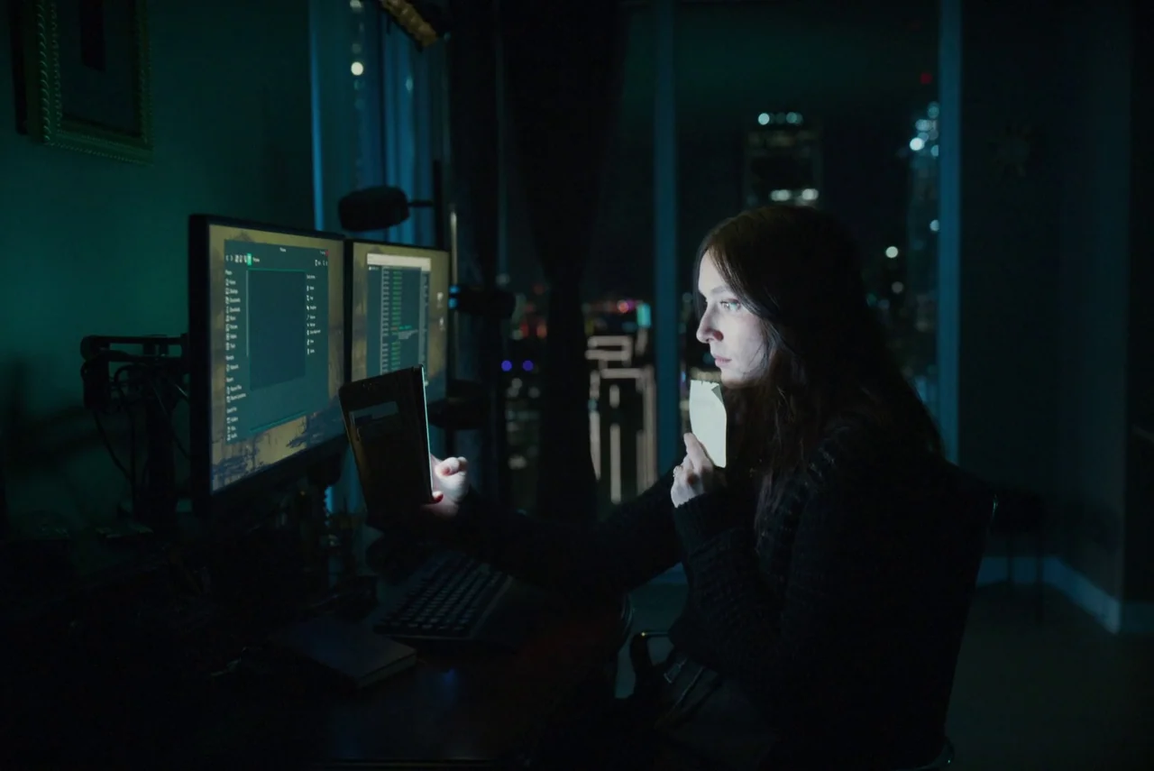 Red Rooms (2023) movie still: medium — A dark, modern office overlooks city lights through a large floor-to-ceiling glass window. A woman sits…