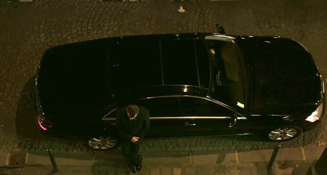 Bastards (2013) movie still: wide — A black sedan is parked along a dark, stone-paved urban curb at night. A man in a dark suit stands leaning…