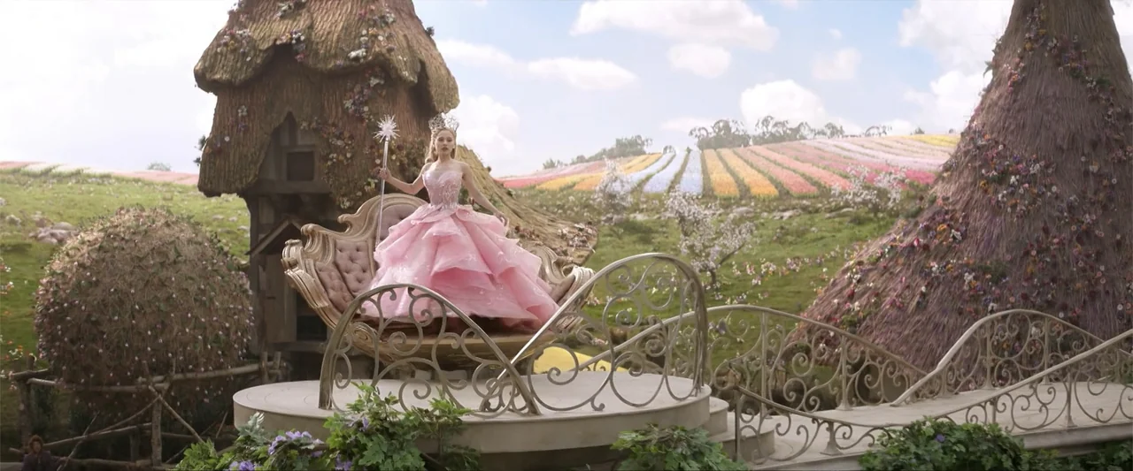 Wicked (2024) movie still: fullbody — A woman stands in a stylized ornate carriage within a vast landscape of colorful flowers. The background…