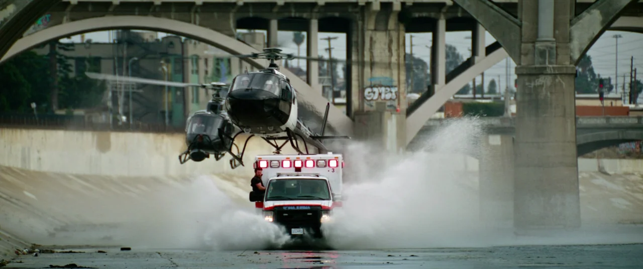 Ambulance (2022) movie still: wide — An ambulance drives rapidly through a wide concrete river canal beneath a large bridge. Two helicopters fly at…