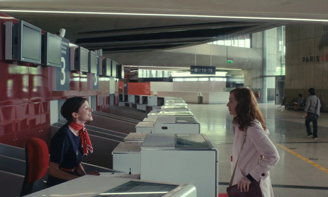 Sidonie in Japan (2023) movie still: medium — An airport check-in hall with sleek red counters and large signage in the background. A traveler…