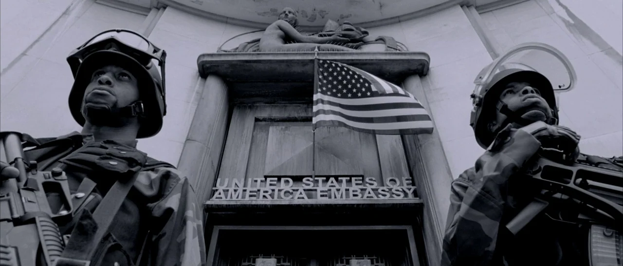 Angel-A (2005) movie still: medium — A stone embassy building entrance features carved relief sculpture above the doorway. Two soldiers in…