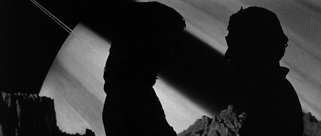 Manhattan (1979) movie still: medium — Two human figures appear in silhouette on a rugged planetary surface. A massive gas giant planet with…