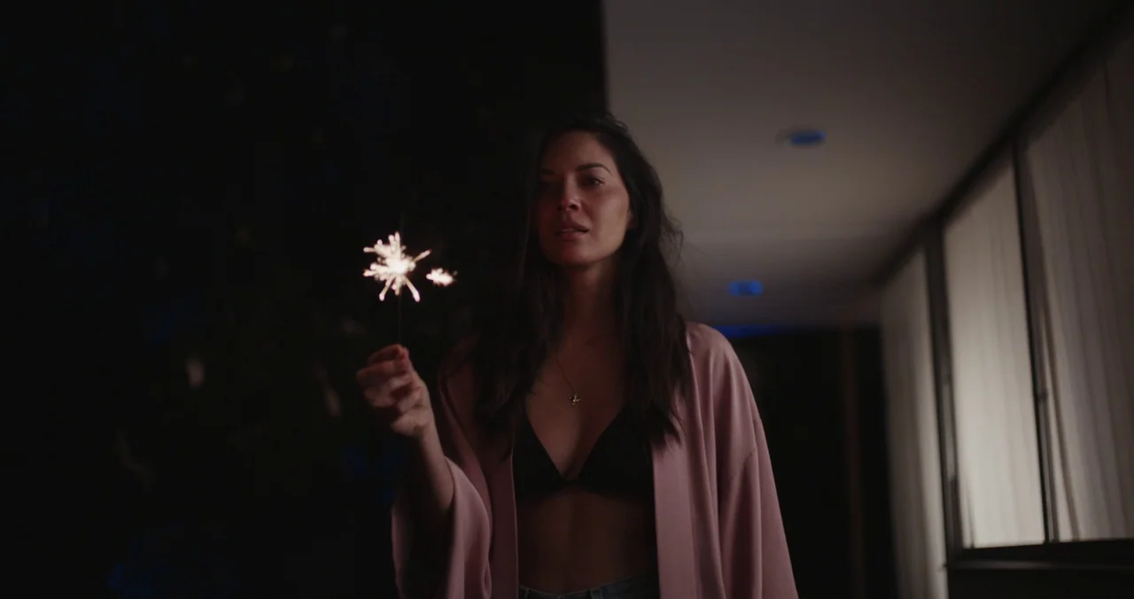 Violet (2021) (2021) movie still: medium — A dimly lit room with a dark wall and a glass window panel. A woman in a pink robe holds a small lit…