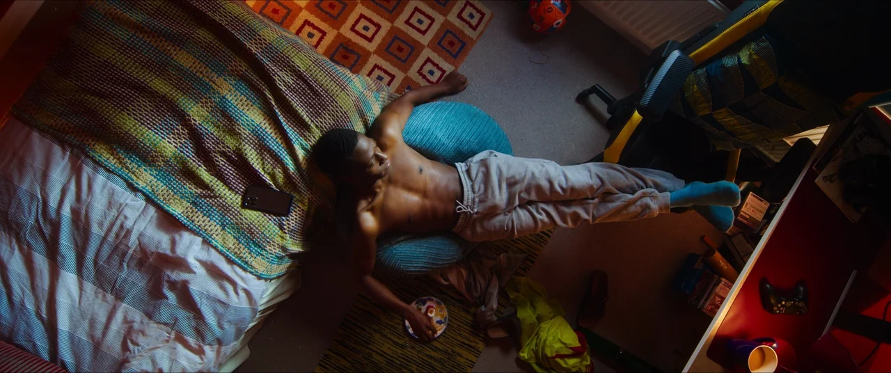 Rye Lane (2023) movie still: wide — A cluttered floor space beside a bed with a patterned quilt and geometric rug. A shirtless young man lies across…