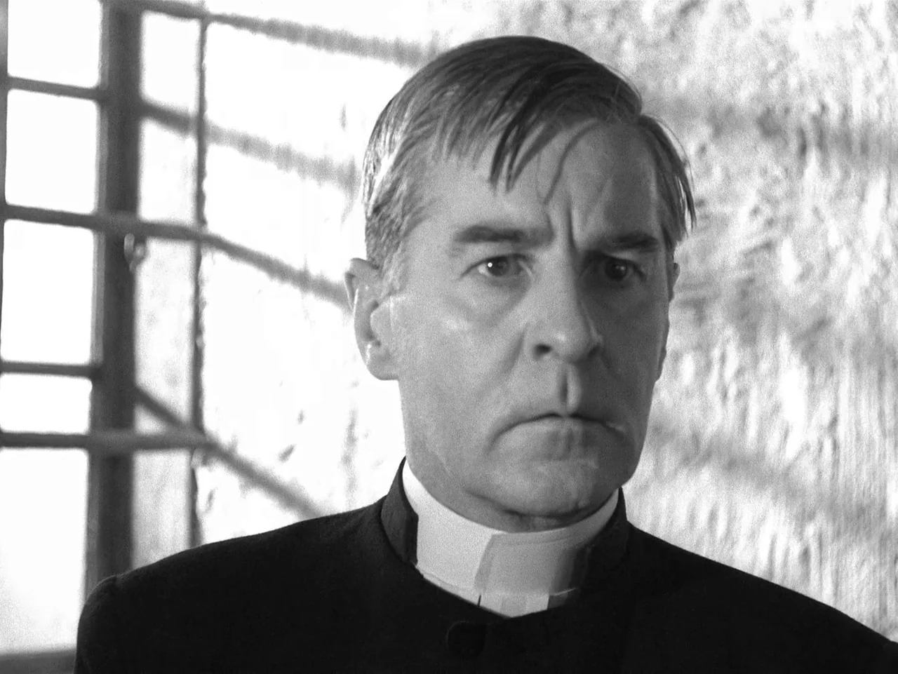 Winter Light (1963) movie still: closeup — A man in clerical attire stands against a textured wall near a window. Sunlight casts grid patterns…