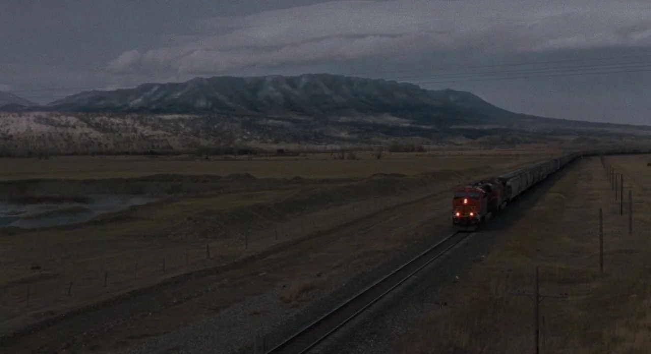 Certain Women (2016) movie still: wide — A long freight train moves through an expansive valley at dusk under heavy clouds. The tracks cut across a…