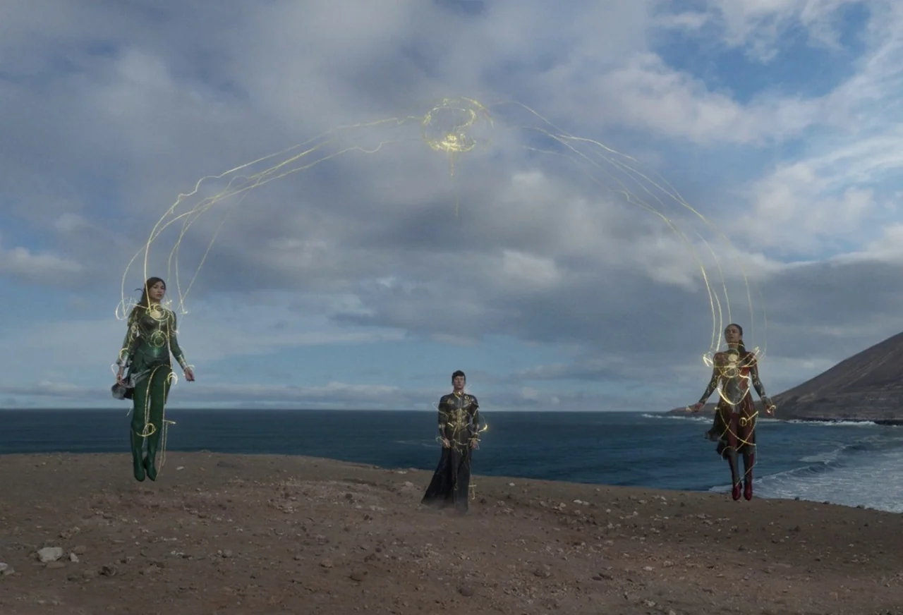 Eternals (2021) movie still: wide — A desolate rocky cliff overlooks a dark blue ocean under a cloudy sky. Three humanoids float above the ground…