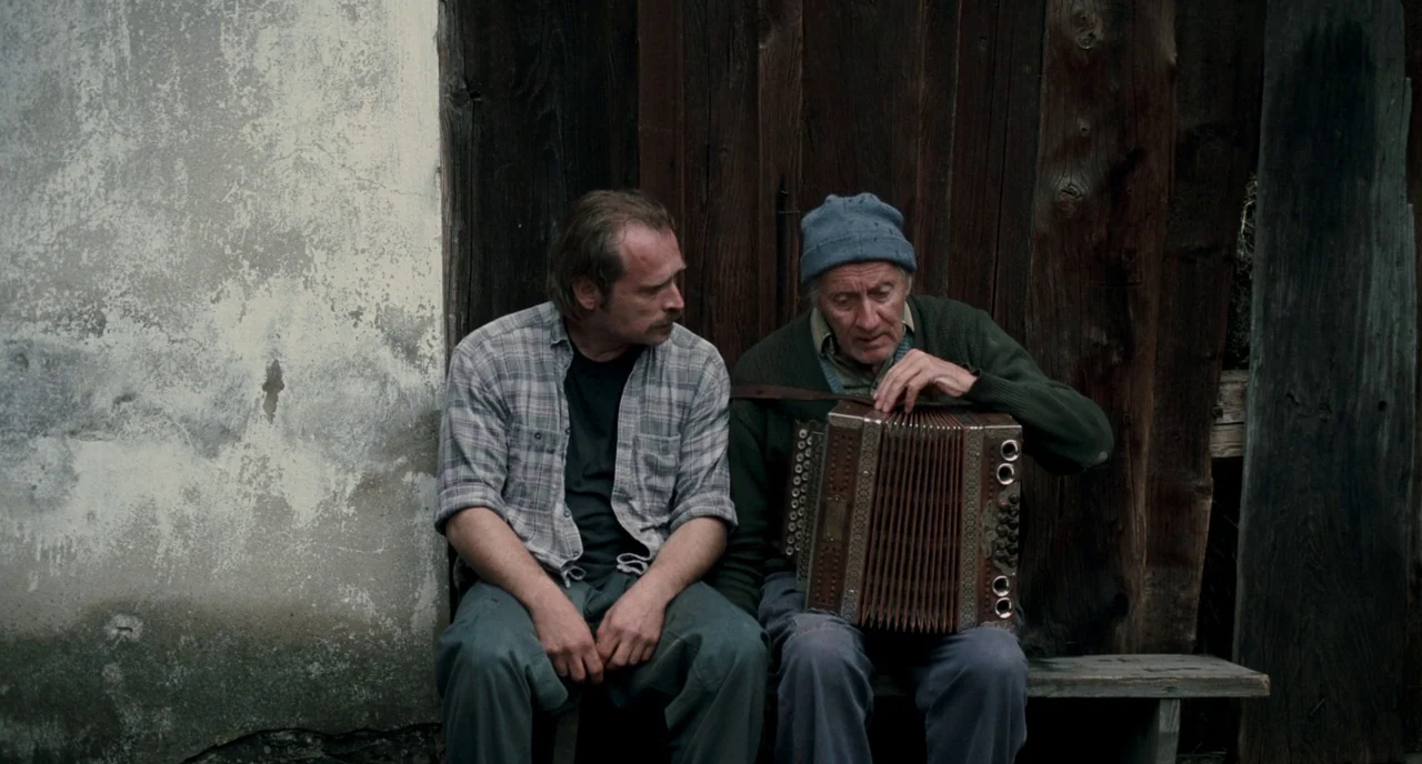 Revanche (2008) movie still: medium — Two men sit side-by-side on a wooden bench against a rustic vertical plank wall. The older man plays a vintage…