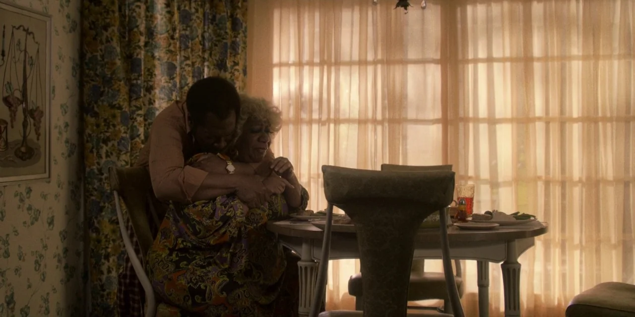 Dolemite Is My Name movie still: medium — A dining room features floral wallpaper and sheer tan curtains covering a large window. A man stands…
