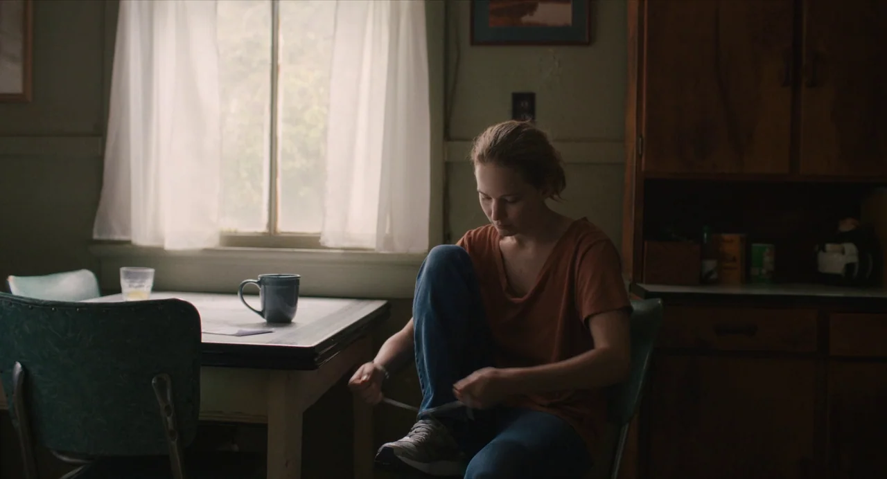 Causeway movie still: medium — A modest kitchen features a small table with a window and white curtains behind. A young woman sits on a chair while…