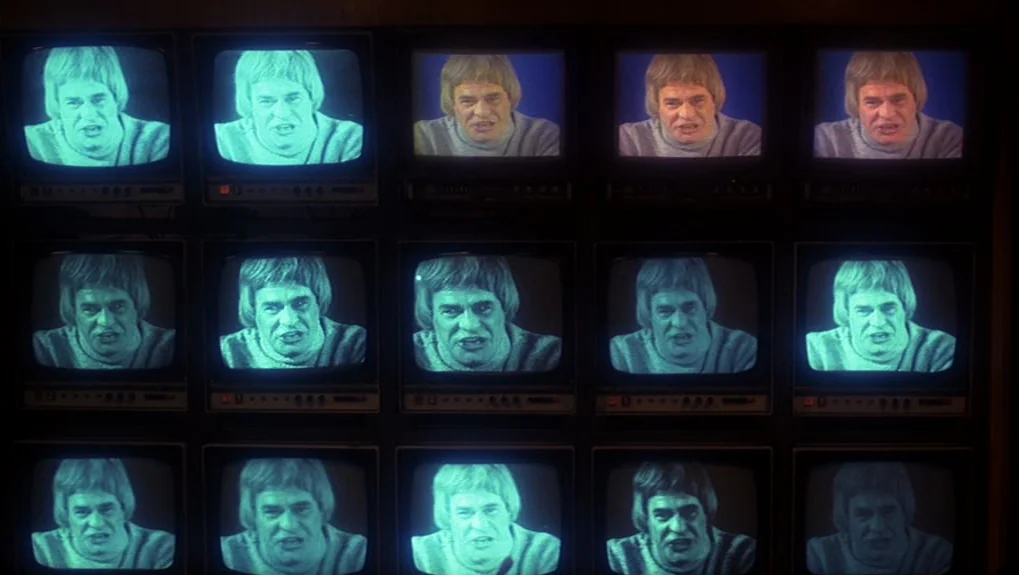 The War of the Worlds: Next Century (1981) movie still: establishing — A dark grid of fifteen identical small television screens mounted on a wall.…
