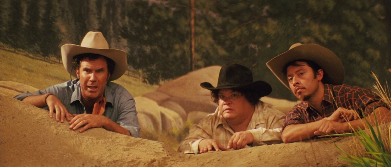 Casa De Mi Padre (2012) movie still: medium — Three men in cowboy hats crouch behind a sandy ridge in front of a painted landscape. They look…