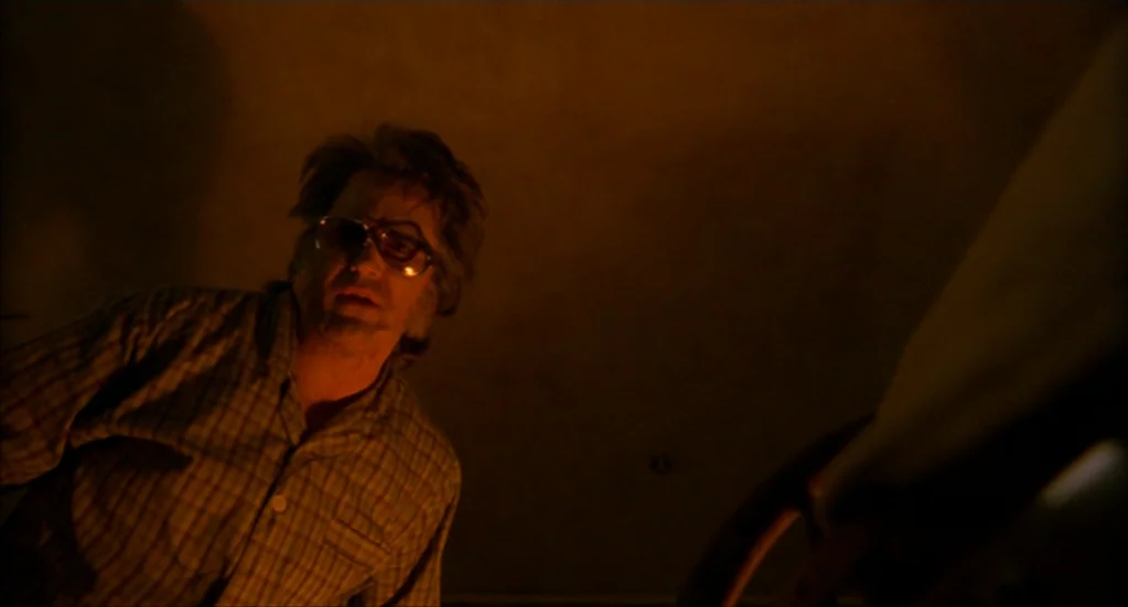 Bubba Ho-Tep (2002) movie still: medium — A dimly lit room is bathed in warm, amber-colored light at night. A middle-aged man looks down towards the…