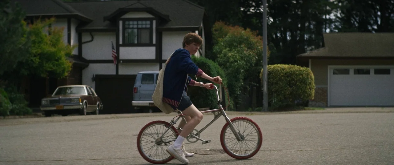 Summer of 84 (2018) movie still: fullbody — A suburban street lined with detached houses features a boy riding a small bicycle. He is moving across…