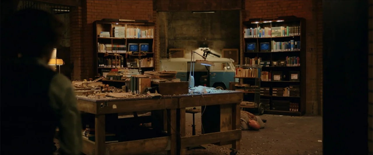 Gunpowder Milkshake (2021) movie still: wide — A brick-walled workspace features a wooden table cluttered with books and a floor-level figure. A…