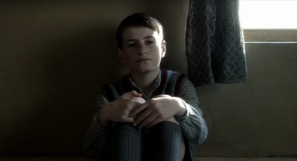 The Long Day Closes (1992) movie still: medium — A dimly lit room with plain walls and a single window to the side. A young boy sits on the floor…