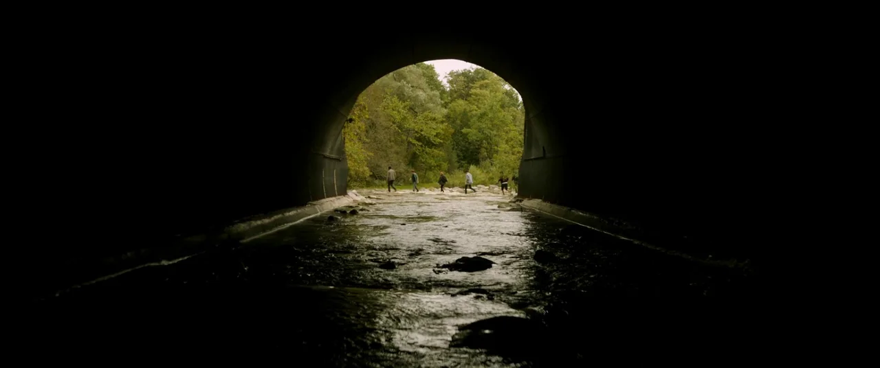 It Chapter Two (2019) movie still: wide — A dark concrete culvert frames a view of a sunlit shallow stream and trees. A group of teenagers walks…