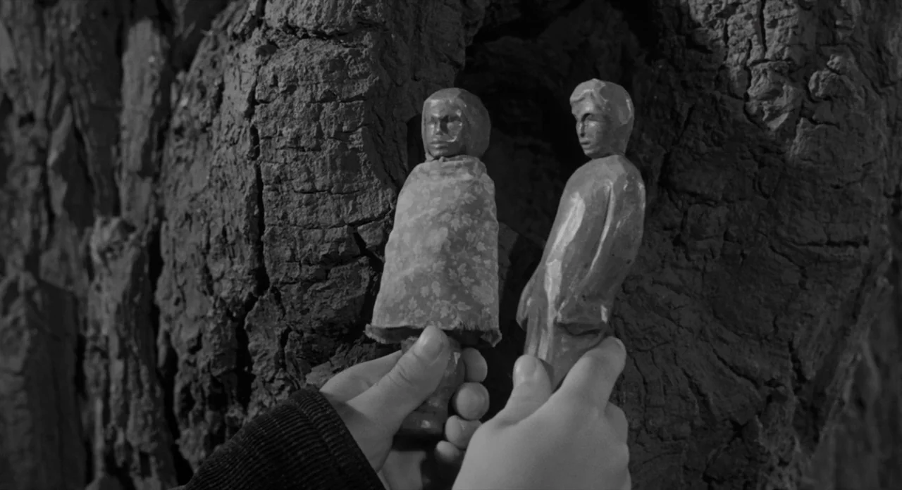 To Kill A Mockingbird (1962) movie still: closeup — A pair of human hands holds two small carved wooden dolls against a tree trunk. The figures…