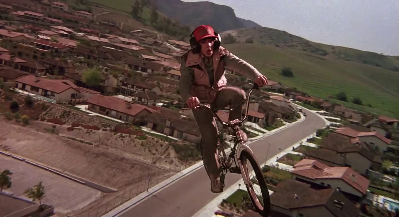 E.T. The Extra-Terrestrial (1982) movie still: fullbody — A suburban residential neighborhood with terracotta-roofed houses sits beneath rolling,…