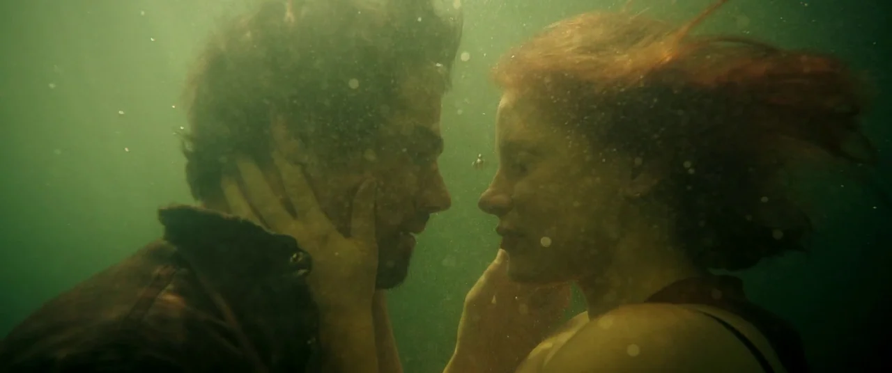 It Chapter Two (2019) movie still: medium — Two people are submerged together in murky, olive-colored water with visible particulate matter. A man…