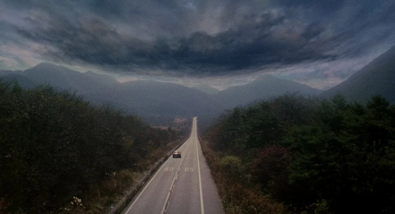 Ringu (1998) movie still: establishing — A long paved road stretches through a valley flanked by dense dark forested hillsides. A massive dark storm…