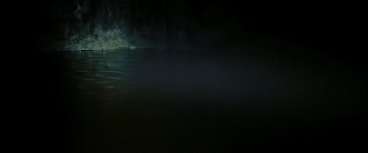 Black Water: Abyss (2020) movie still: establishing — A dark subterranean cavern contains a body of still, reflective water. The stone wall curves…
